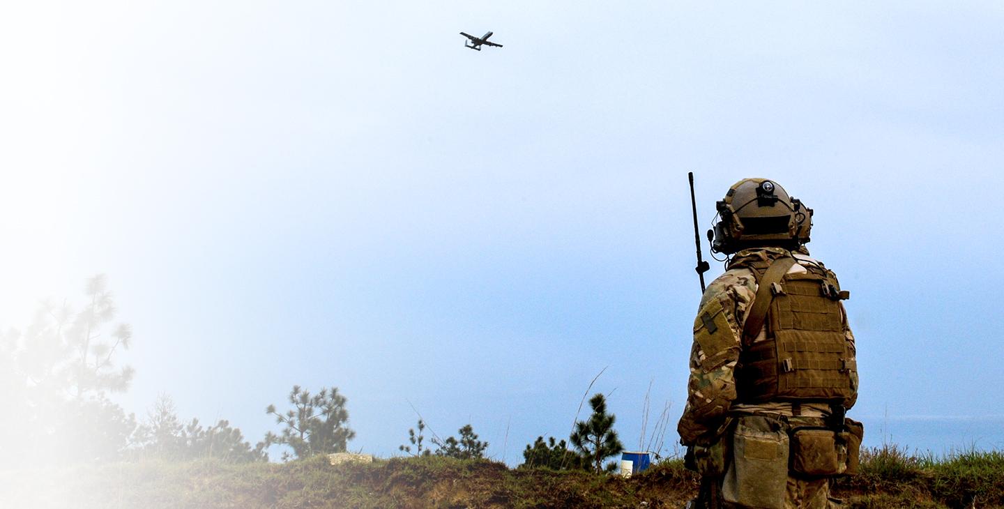 JTAC warfighter  in the field holding a military satcom antenna looking up at an aircraft flying by